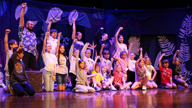 Students gathered on a theater stage, some standing and some kneeling, each with their right first raised in the air at the end of a production.