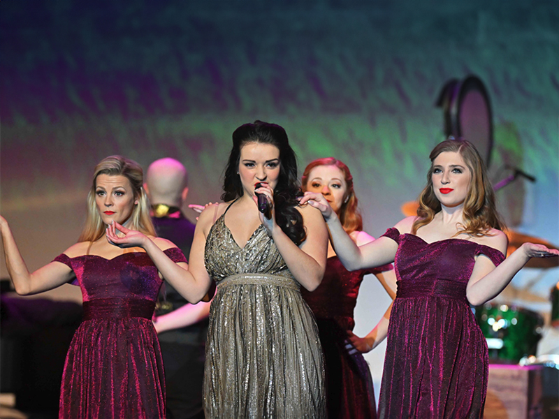 Five women in white dresses and gold tiaras stand on a stage singing