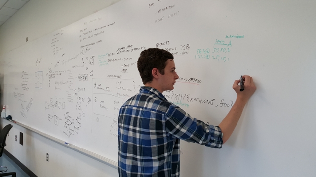 A male student writes on dry erase board with many equations