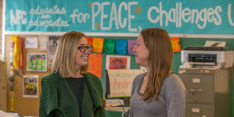two people talking in a classroom with a peace poster in the background