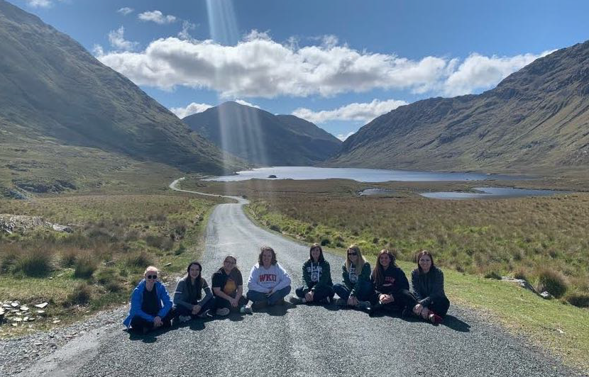 a group of students sitting on a road in a single line with a view of a mountain range behind them