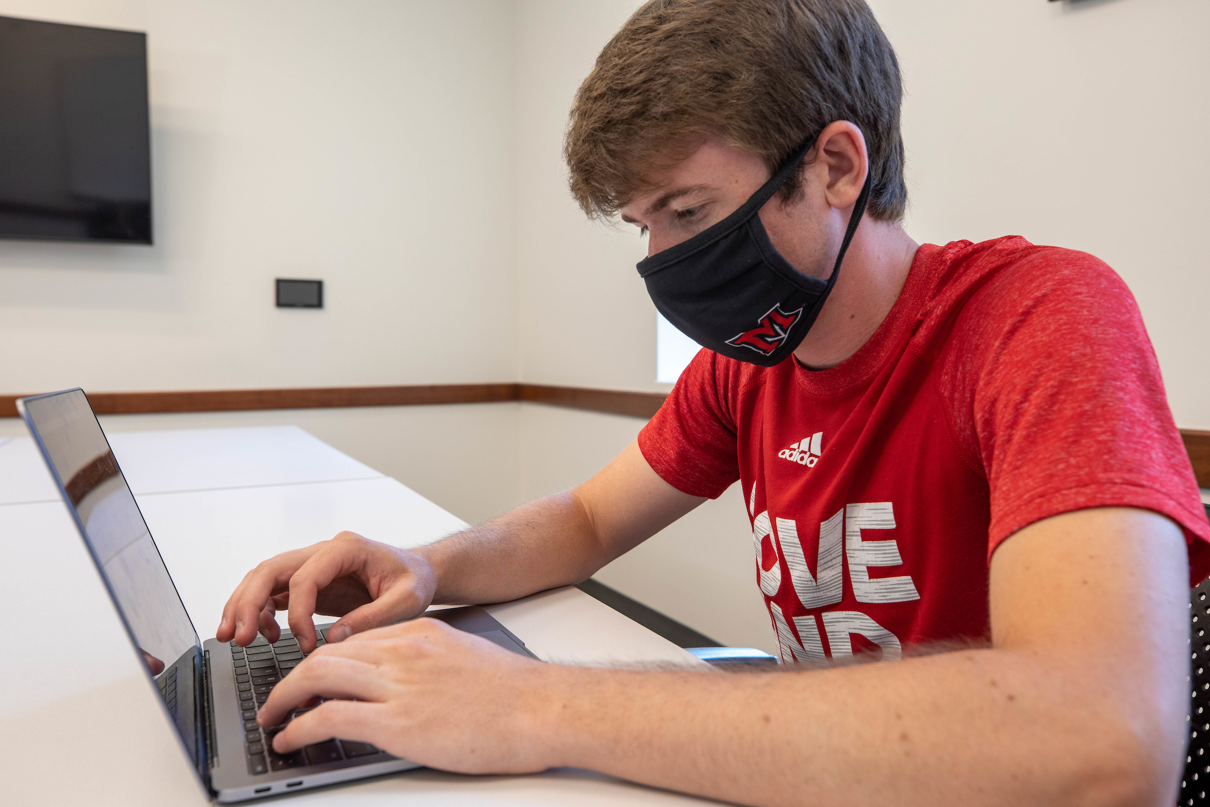 Brown-haired boy wearing a black mask and red Miami shirt sits at a long white table typing on a laptop.