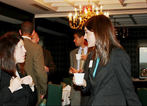 Alumni chatting at a previous networking breakfast. There are two women in the foreground, the one on the right is holding a drink. Two men in the background. All are wearing business attire.