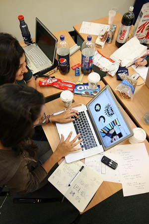 Two female students sit around a laptop computer working on a presentation or project. The table is covered with drinks and junk food wrappers.