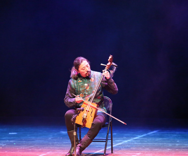  A man sits along on stage playing a string instrument that he holds in his lap