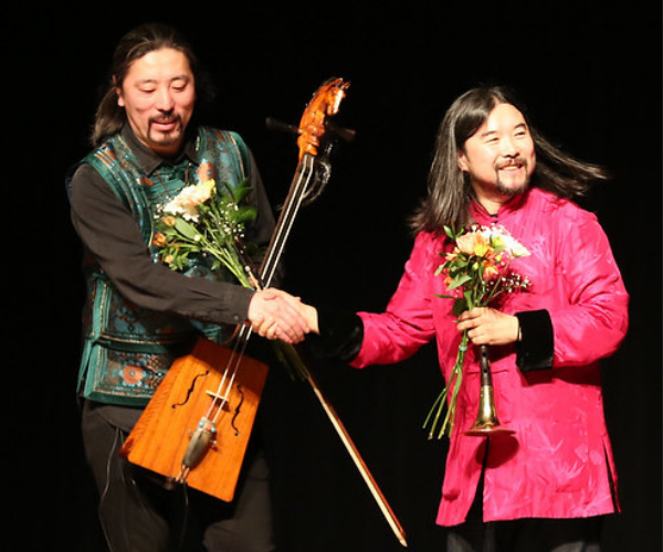  two performers shake hands. one of them holds their instrument while the other holds flowers