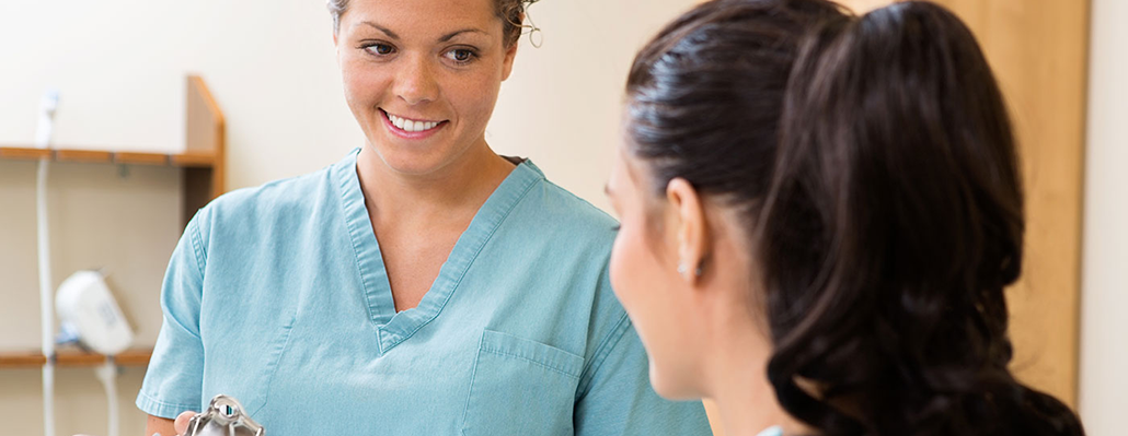 A woman in scrubs holds a clipboard. She smiles while facing another person