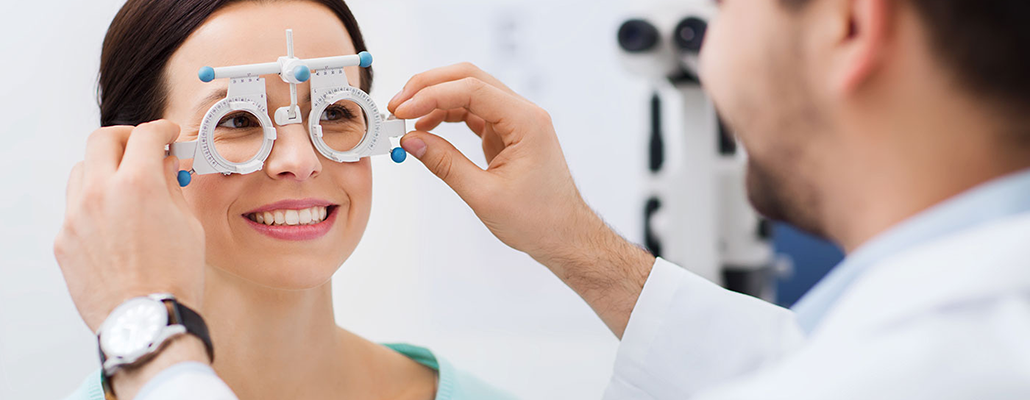 A smiling woman is being evaluated with optician's equipment. The optician seen opposite her adjusts the equipment.