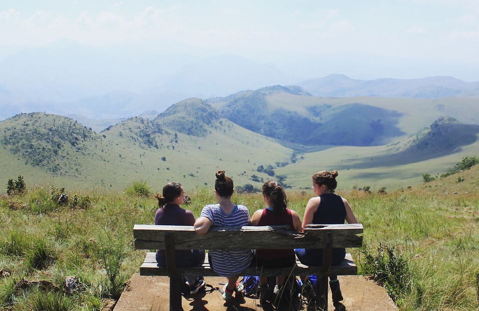 students sit on a bench, facing away from camera, and gaze out at distant mountains