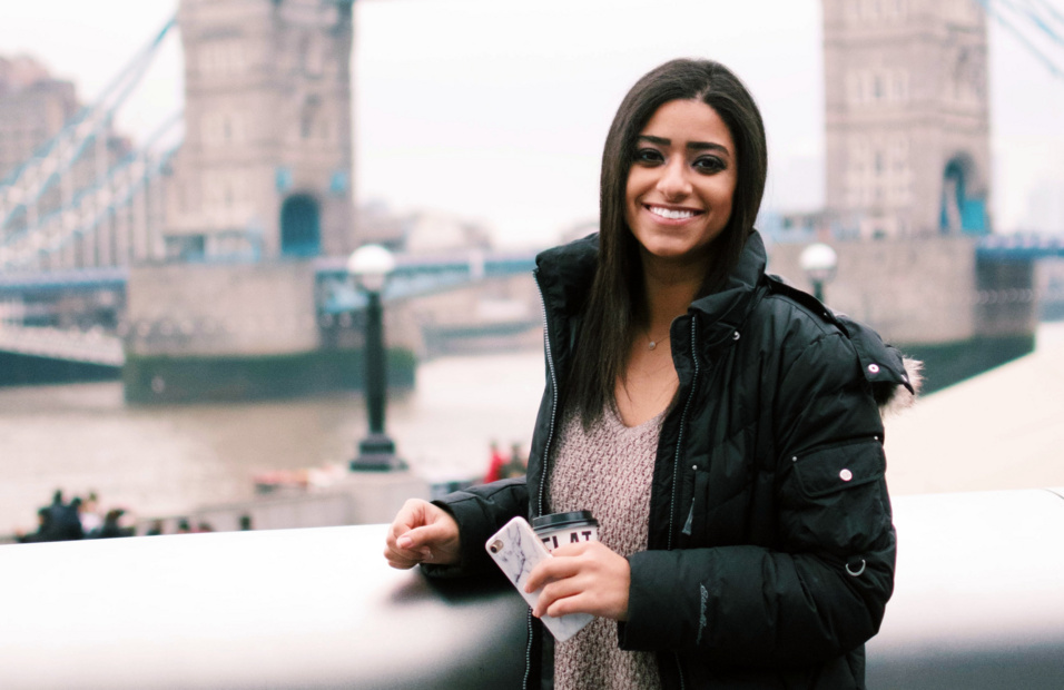 A young woman smiles at the camera. In the background is London Bridge