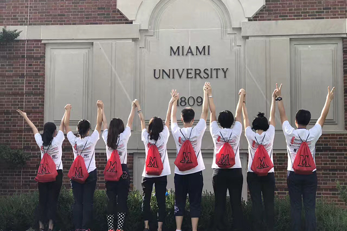Orientation students wearing red backpacks face away from the camera in front of Miami University 1809 sign. They hold hands and raise them high