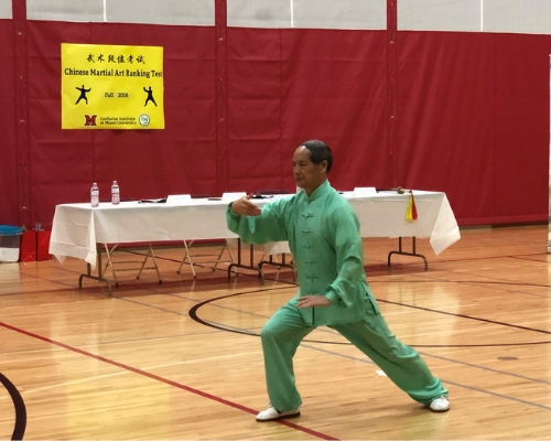 One of the judges performs tai chi for the audience after the students take their test. He is dressed in a green robe as he lunges forward in a tai chi move. 