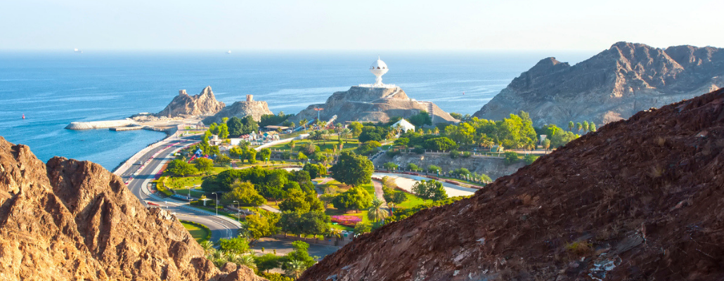 a picture of the countryside in Oman. There is a mountain in the foreground with the sea and buildings in the back
