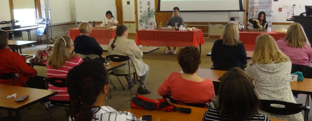 many staff watching a student pour tea during a tea ceremony at the front of MacMillan Hall's great room, all are sitting in chairs and tables