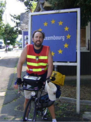 a middle-aged man wears an orange hazard vest and sits on a bicycle in front of a sign that says Luxembourg