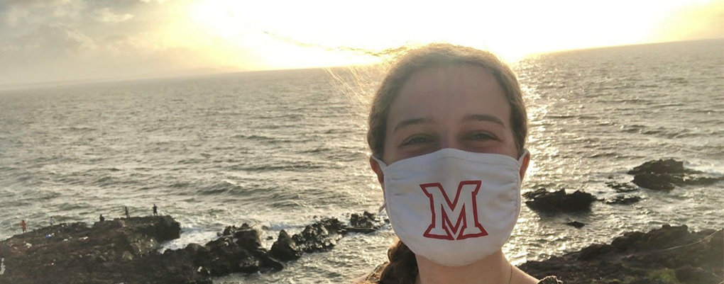 Anna, wearing a face mask, poses by the seashore