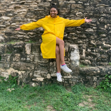 Arianna is dressed in a bright yellow coat and stretches out her arms against a stone structure in Belize