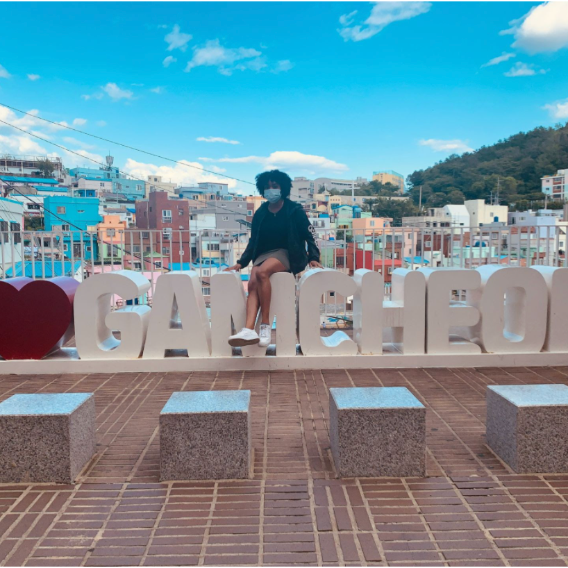 Meagan poses on a sign that reads I heart Gamicheon, with city in background