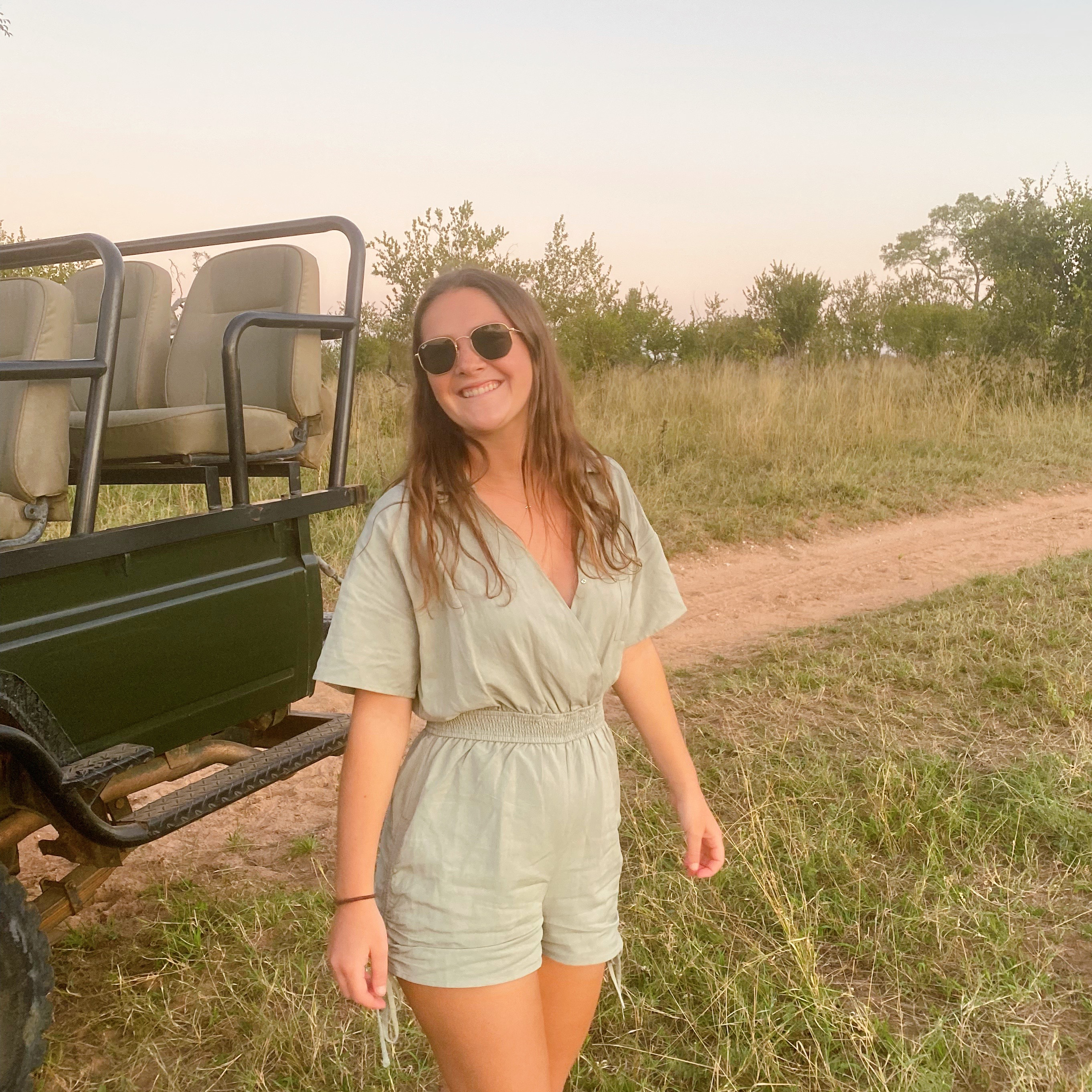 Jessica smiles at the camera as she stands in a grassy field in South Africa. An open vehicle with seating on top is visible in the background.