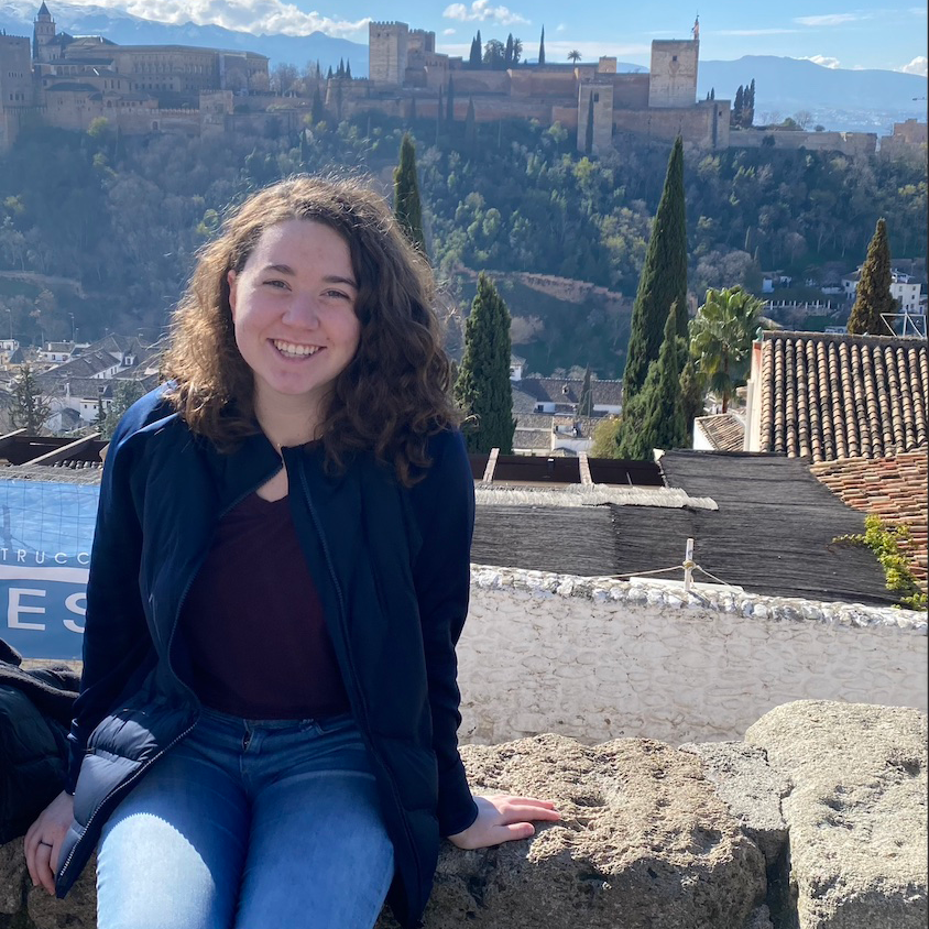 Katherine sits on a stone wall. Behind her is the city of Valencia, including picturesque castle