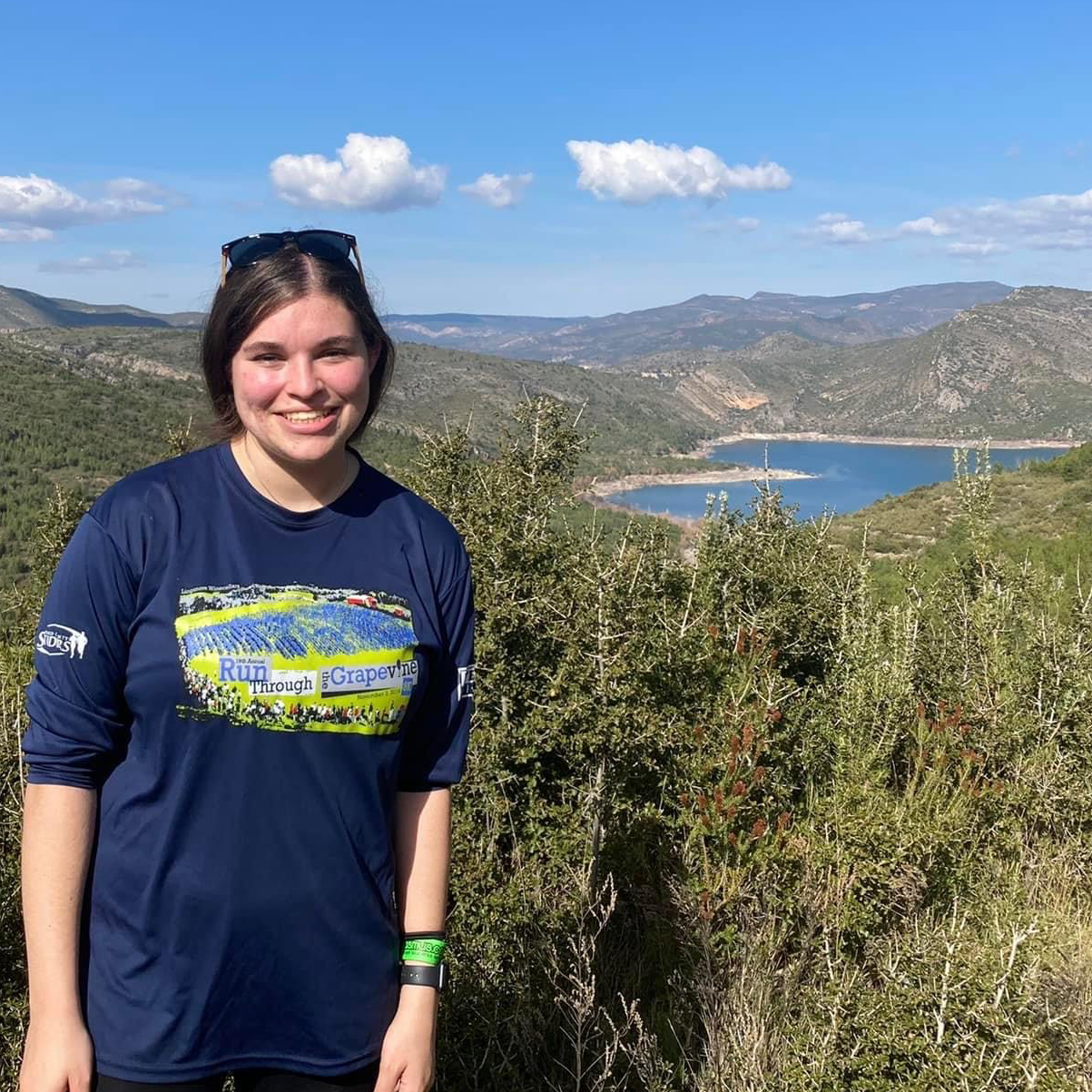 Ellie smiles at the camera as she stands in an outdoor setting in Spain. A body of water and rolling hills are visible in the background