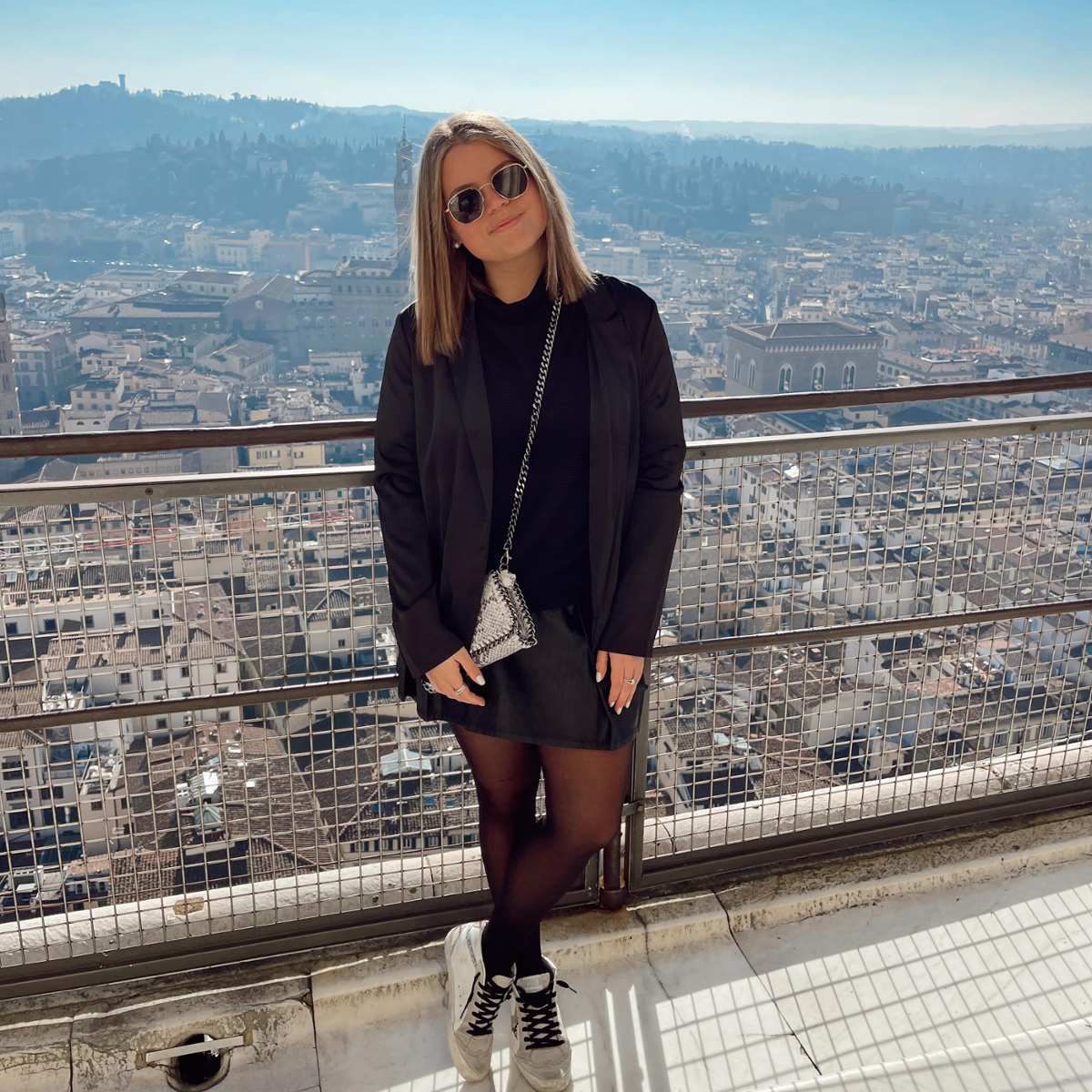 Samantha, wearing sunglasses, poses on a walkway. Distant mountains or hills and the buildings of the city of Florence are visible in the background