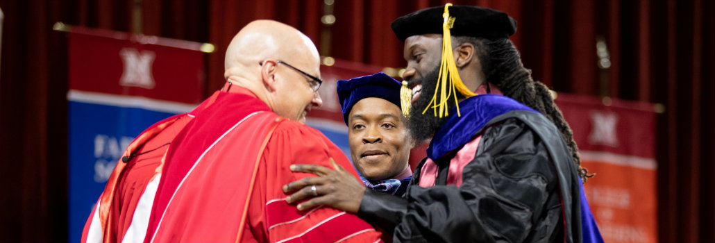 Picture of a doctoral student with their hooding professor, shaking hands with President Crawford at December graduation