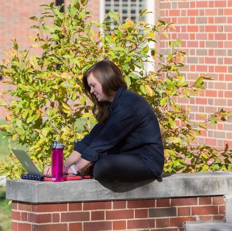 A female student sitting on a ledge outside of her residence hall working on a laptop