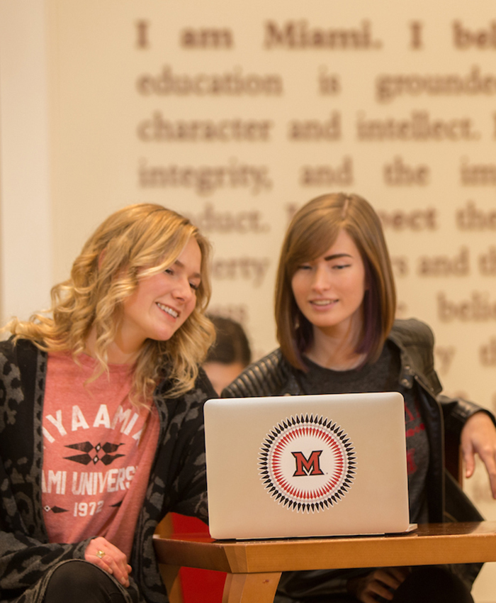 Two students sit in Armstrong Student Center, looking at a computer with a decal on its cover. A Miami beveled M is encircled by ribbonwork pattern on the decal.