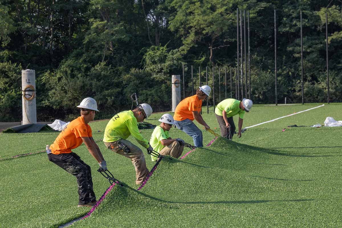 Several workers pull on large hooks grabbing the synthetic turf at Chestnut Fields to pull it smooth