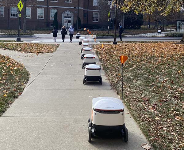 Five Starship food delivery robots in a line on the sidewalk between Armstrong Student Center and Irvin Hall, near the Spring Street crosswalk