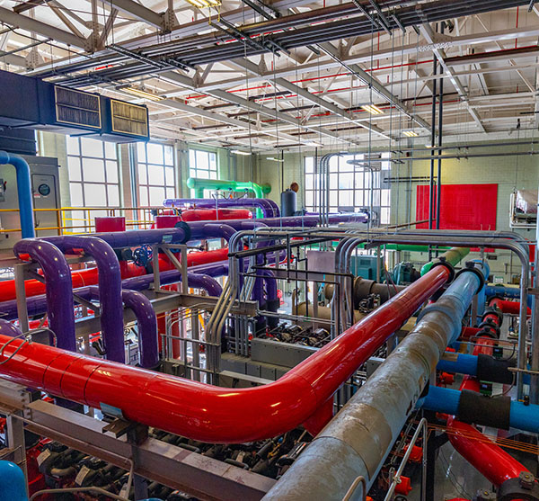 Inside view of geothermal and heating hot water pipes in Billings Hall, looking from above. Large windows are to the left and the old natatorium scoreboard can be seen in the background on the wall