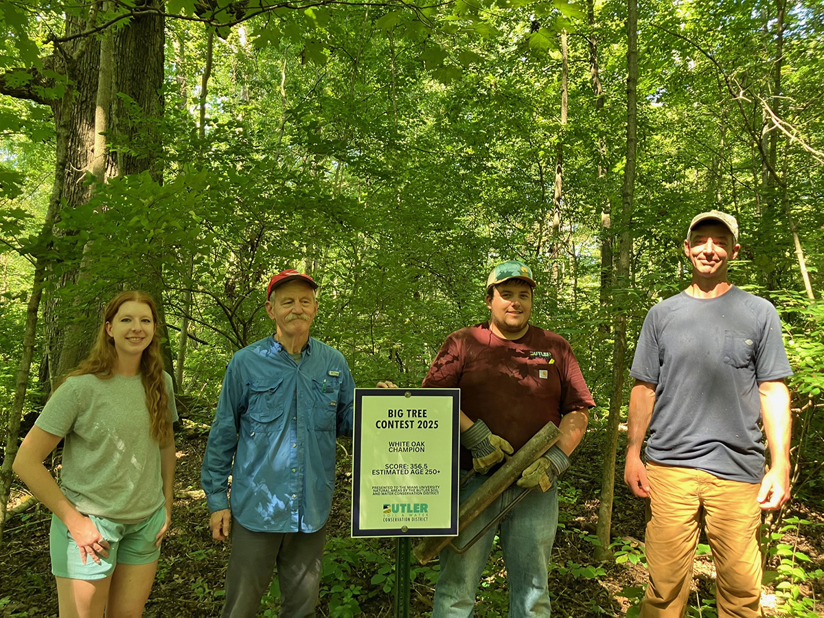 Ashlee Mulrooney, Steve Gordon, Brady Smith, and Rob Atkinson pose under the big white oak and behind a sign indicating the dimensions of the Big Tree Contest winner