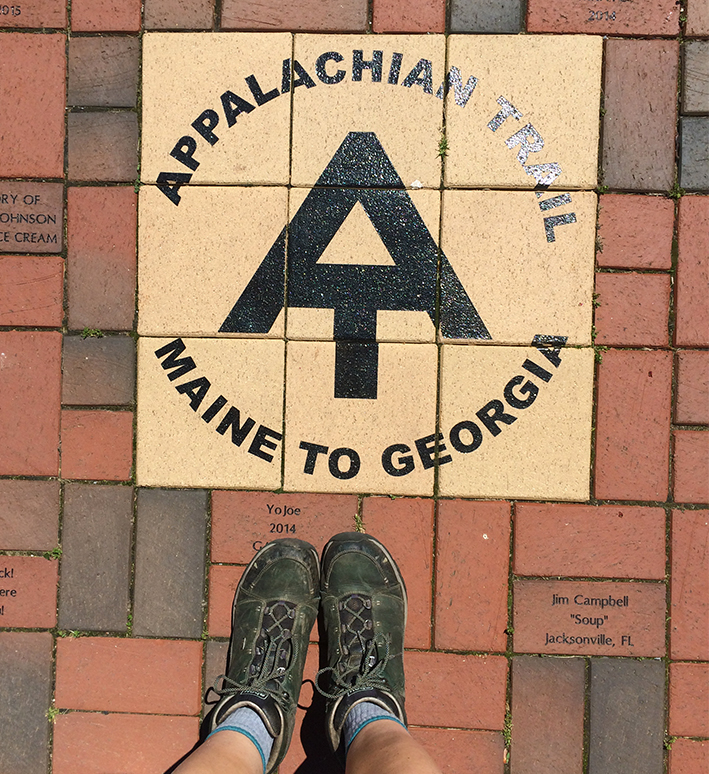 Tess's feet, in hiking boots, stand by a plaque on a path that says 'Appalachian Trail Maine to Georgia'