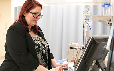 A female standing a computer with a hospital bed in the background. 