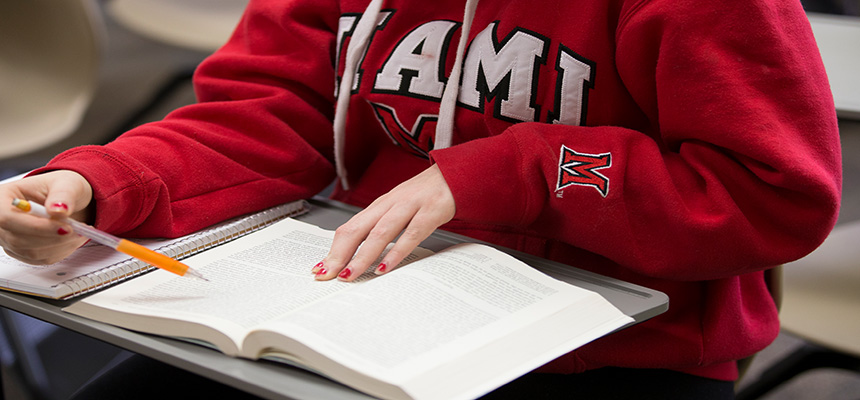  Image of a textbook on a desk with a pencil in someones hand and a Miami red sweatshirt