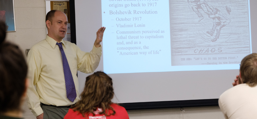 Matthew Smith standing in front of the classroom teaching with a piece of paper in his hand. 