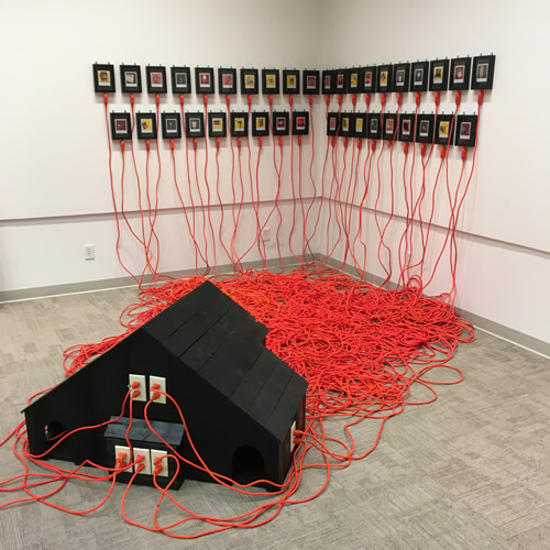 Installation on floor and wall in corner of gallery. In foreground a black wooden box shaped similar to a tobacco barn has cream-colored power outlets. Each outlet has an orange plug with orange cables that run toward the corner. On the wall are two rows of black-framed images, 20 on the top row and 18 on the bottom row.