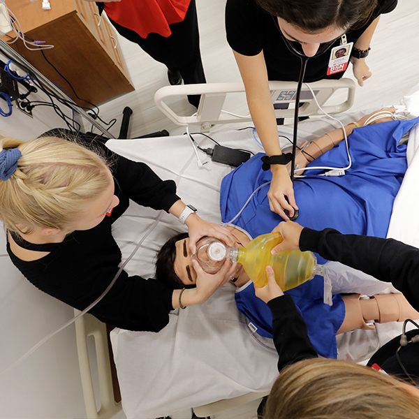  3 students working on the simulation manikin by checking his heart, giving him oxygen, and checking his blood pressure.