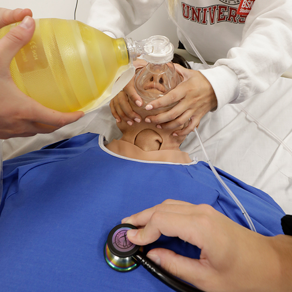  A student holding oxygen over the simulation manikin's mouth and another checking his heart with a stethoscope. 