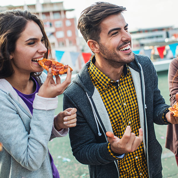 2 people laughing outside eating slices of pizza.