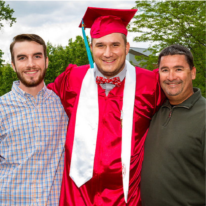A male in his graduation cap and gown his is arms around 2 other men during commencement on the Middletown campus. 