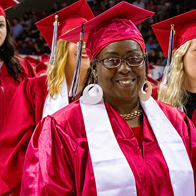A female at commencement in her cap and gown smiling at the camera before walking up to receive her diploma.