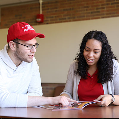An admission counselor talking to a prospective student while looking at a book on a table. 