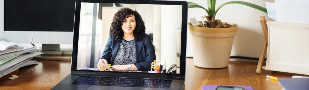 Organized desk with lap top open and on the screen appears a person smiling and wearing a suit