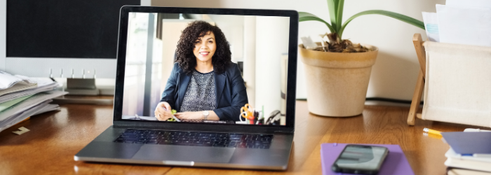 Organized desk with lap top open and on the screen appears a person smiling and wearing a suit