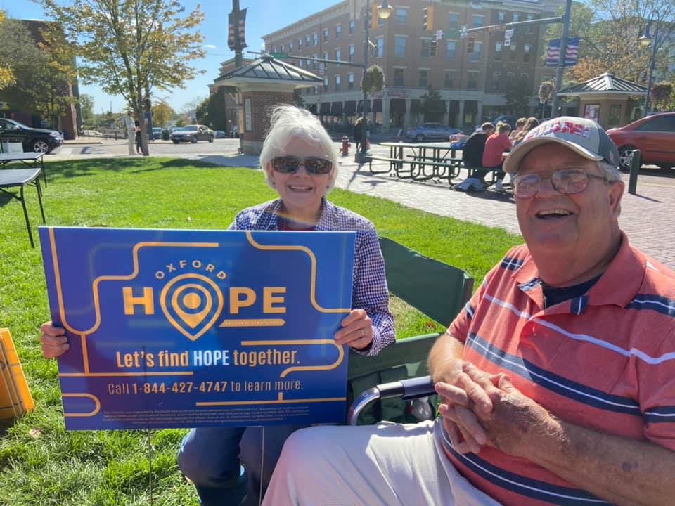 Two senior Oxford community members holding a yard sign uptown that reads Oxford Hope. Let's find HOPE together. 
