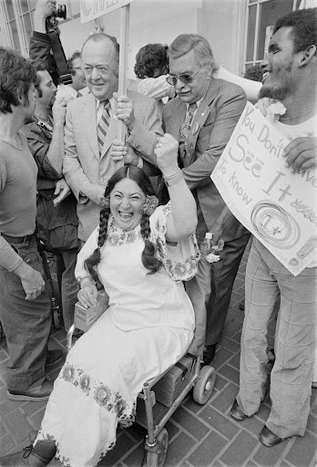 black and white image from 1977 protests. A woman in long braids and a dress sits in a wheelchair with her hand raised in triumph. A group of men stand behind her holding signs. 