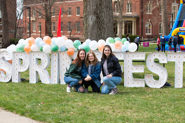 Huge marquee letters spelling spring fest outside on the academic quad, with a few students crouched in front. 
