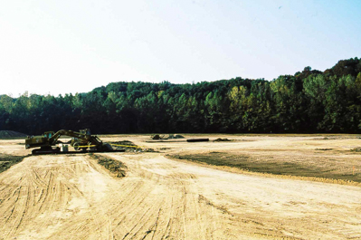 A field of dirt compressed by tire tracks surrounded by a forest